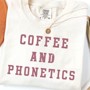 May include: Ivory-colored t-shirt with the words "COFFEE AND PHONETICS" in a maroon, distressed font. A gold necklace with a pendant is draped across the shirt. A brown hat is visible on the left side.