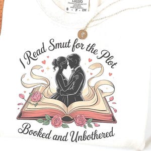 May include: Ivory-colored t-shirt with the text "I Read Smut for the Plot" and "Booked and Unbothered." The graphic features a couple embracing over an open book, with roses and ribbons. A gold necklace is draped over the shirt.