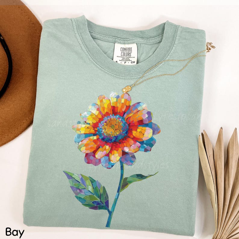以下が含まれることがあります： セージグリーンの半袖Tシャツで、首元にComfort Colorsのラベルが付いています。前面には、オレンジ、黄色、青、紫の花びら、緑の茎、一枚の葉で構成された、鮮やかなマルチカラーのモザイクスタイルの花のグラフィックが描かれています。