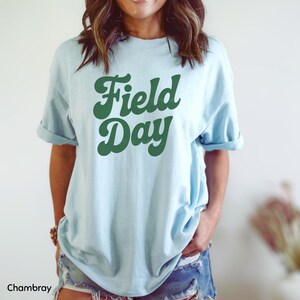 May include: Light blue t-shirt with the words "Field Day" in a retro green font. The shirt has rolled-up sleeves and is made of a soft material. The word "Chambray" is at the bottom left of the image.