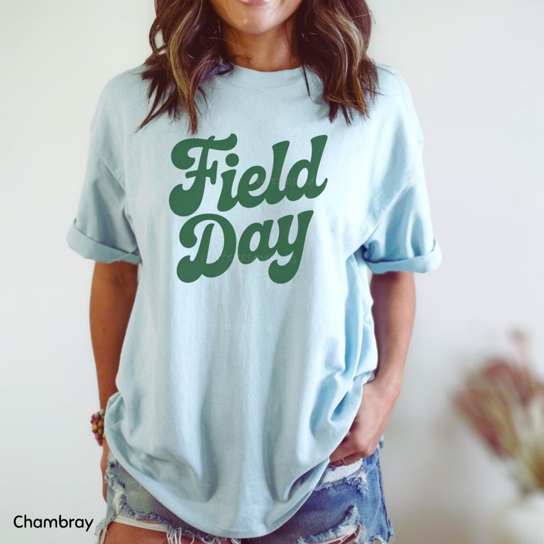 May include: Light blue t-shirt with the words "Field Day" in a retro green font. The shirt has rolled-up sleeves and is made of a soft material. The word "Chambray" is at the bottom left of the image.