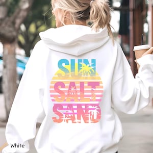 K&ouml;nnte beinhalten: Ein wei&szlig;er Kapuzenpullover mit einer Grafik auf der R&uuml;ckseite und dem Text SUN SALT SAND. Das Design zeigt ein kreisf&ouml;rmiges Sonnenuntergangsmotiv in gelben, orangen und rosa Verl&auml;ufen, &uuml;berlagert mit Palmensilhouetten und horizontalen Linien.