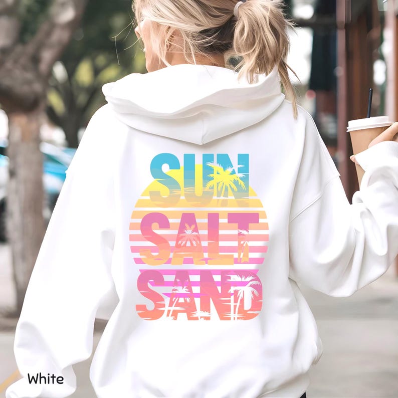 K&ouml;nnte beinhalten: Ein wei&szlig;er Kapuzenpullover mit einer Grafik auf der R&uuml;ckseite und dem Text SUN SALT SAND. Das Design zeigt ein kreisf&ouml;rmiges Sonnenuntergangsmotiv in gelben, orangen und rosa Verl&auml;ufen, &uuml;berlagert mit Palmensilhouetten und horizontalen Linien.
