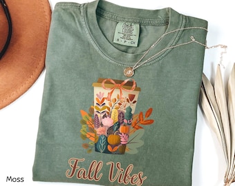 May include: A moss green t-shirt featuring a graphic of a coffee cup filled with colorful autumn leaves and pumpkins, topped with an orange bow. The text "Fall Vibes" is printed in a cursive font below the graphic. The shirt has a crew neckline.