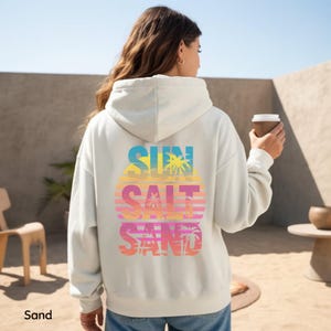 K&ouml;nnte beinhalten: Ein sandfarbener Kapuzenpullover mit einer gro&szlig;en Grafik auf der R&uuml;ckseite, die den Text SUN SALT SAND in fetten Buchstaben zeigt. Das Design enth&auml;lt ein Sonnenuntergangsmotiv mit horizontalen Streifen und Palmensilhouetten in Gelb, Pink und Blau.