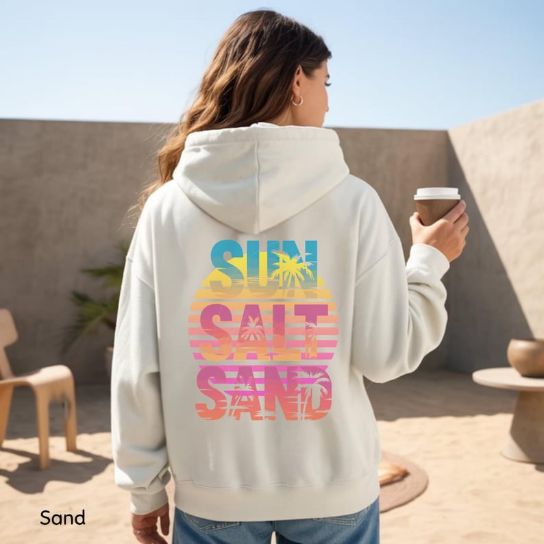 K&ouml;nnte beinhalten: Ein sandfarbener Kapuzenpullover mit einer gro&szlig;en Grafik auf der R&uuml;ckseite, die den Text SUN SALT SAND in fetten Buchstaben zeigt. Das Design enth&auml;lt ein Sonnenuntergangsmotiv mit horizontalen Streifen und Palmensilhouetten in Gelb, Pink und Blau.