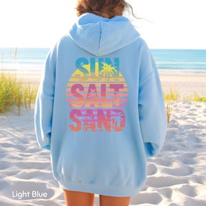 K&ouml;nnte beinhalten: Ein hellblauer Kapuzenpullover mit einer Grafik auf der R&uuml;ckseite, die den Text SUN SALT SAND in einem Farbverlauf aus Blau, Gelb, Orange und Pink zeigt. Das Design enth&auml;lt ein Sonnenuntergangsmotiv mit Palmensilhouetten &uuml;ber dem Text.