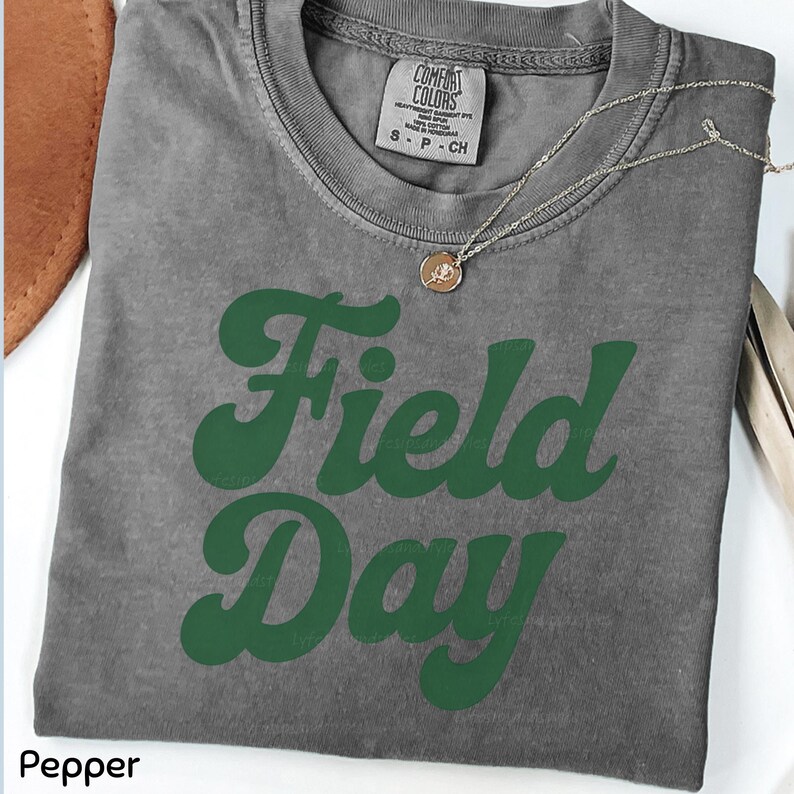 May include: A gray t-shirt with the words "Field Day" in green, retro-style lettering. The shirt has a crew neck and short sleeves. A gold necklace with a pendant is draped across the shirt. The shirt is folded.