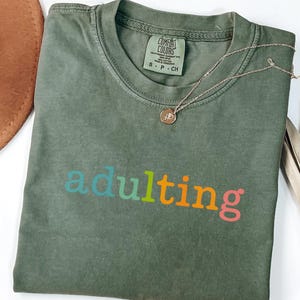 May include: A moss green t-shirt with the word "adulting" in colorful letters. The shirt has a crew neck and short sleeves. A gold necklace with a small pendant is draped over the shirt.