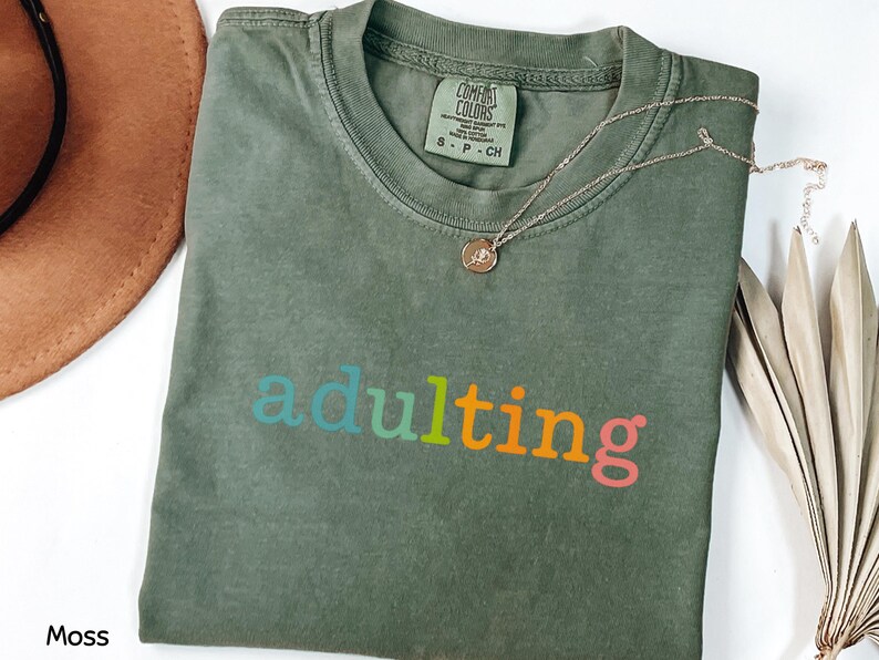 May include: A moss green t-shirt with the word "adulting" in colorful letters. The shirt has a crew neck and short sleeves. A gold necklace with a small pendant is draped over the shirt.