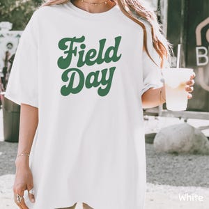 May include: White t-shirt with the words "Field Day" in a green, retro font. The shirt has short sleeves and a relaxed fit. The image shows the shirt being worn.