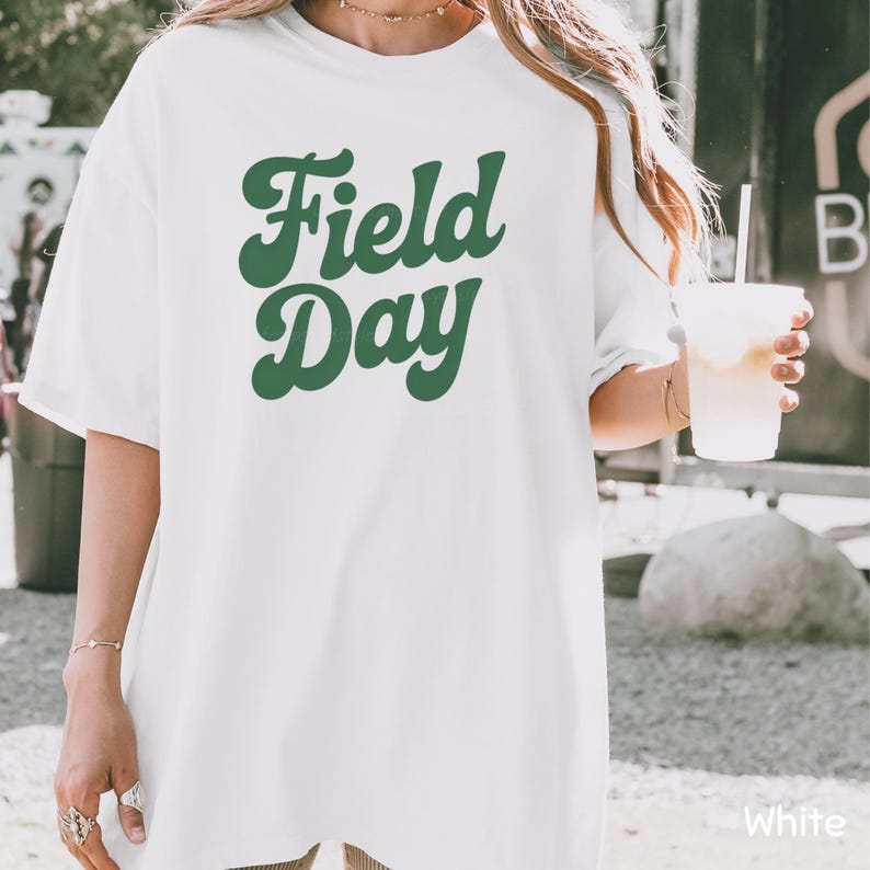 May include: White t-shirt with the words "Field Day" in a green, retro font. The shirt has short sleeves and a relaxed fit. The image shows the shirt being worn.