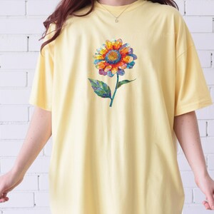 以下が含まれることがあります： 淡い黄色の半袖Tシャツ。中央には、赤、オレンジ、青、紫の色合いでモザイク調に描かれた花びらを持つ一輪の花のグラフィックがプリントされており、緑色の茎と葉が添えられています。