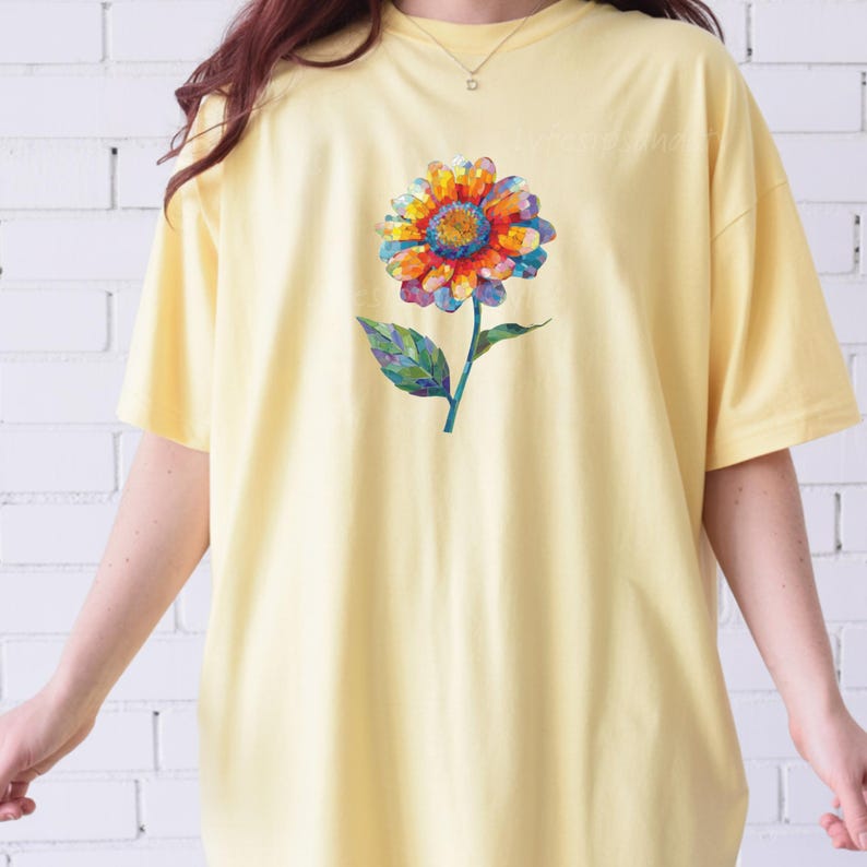 以下が含まれることがあります： 淡い黄色の半袖Tシャツ。中央には、赤、オレンジ、青、紫の色合いでモザイク調に描かれた花びらを持つ一輪の花のグラフィックがプリントされており、緑色の茎と葉が添えられています。