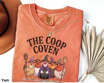 May include: A burnt orange short-sleeve t-shirt featuring the text THE COOP COVEN in black letters above a graphic of five chickens wearing colorful witch hats and capes, with some holding broomsticks. The shirt has a crew neckline and a tag.