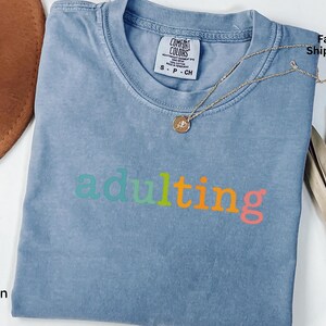 May include: Blue jean colored t-shirt with the word "adulting" in a rainbow of colors. A gold necklace with a pendant rests on the shirt. The shirt is folded and has a Comfort Colors tag.