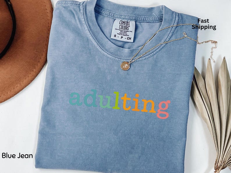 May include: Blue jean colored t-shirt with the word "adulting" in a rainbow of colors. A gold necklace with a pendant rests on the shirt. The shirt is folded and has a Comfort Colors tag.