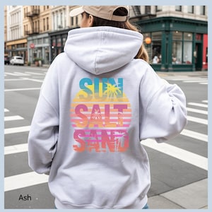 K&ouml;nnte beinhalten: Ein aschfarbener Kapuzenpullover mit einem gro&szlig;en Grafikdruck auf der R&uuml;ckseite. Das Design zeigt ein kreisf&ouml;rmiges Sonnenuntergangsmotiv in gelben, orangen und rosafarbenen Verl&auml;ufen mit Palmensilhouetten und dem Text SUN SALT SAND in Blockschrift.