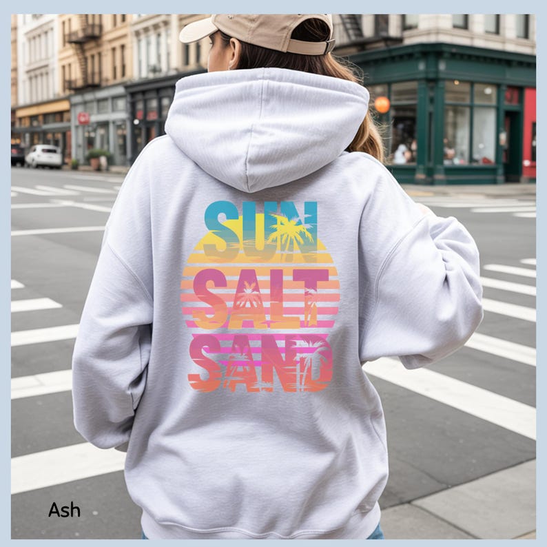 K&ouml;nnte beinhalten: Ein aschfarbener Kapuzenpullover mit einem gro&szlig;en Grafikdruck auf der R&uuml;ckseite. Das Design zeigt ein kreisf&ouml;rmiges Sonnenuntergangsmotiv in gelben, orangen und rosafarbenen Verl&auml;ufen mit Palmensilhouetten und dem Text SUN SALT SAND in Blockschrift.