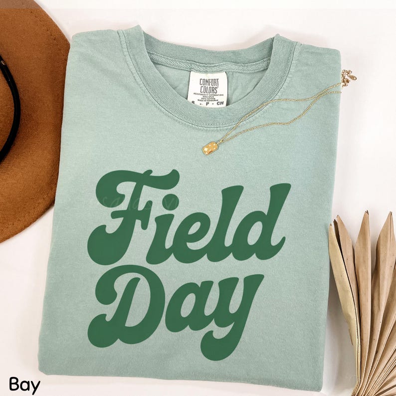 May include: A light green t-shirt with the words "Field Day" in large, dark green, retro-style lettering. A gold necklace with a small pendant rests on the shirt. A brown hat and dried leaves are partially visible.