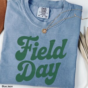 May include: A blue jean Comfort Colors t-shirt with the words "Field Day" in green, retro-style lettering. A gold necklace with a small pendant is draped across the shirt. The shirt is size small.