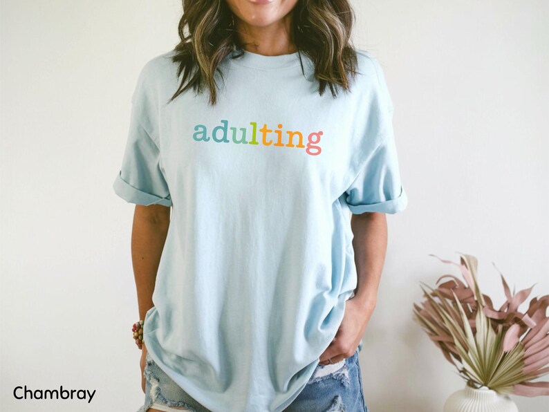 May include: Light blue t-shirt with the word "adulting" in rainbow colors. The shirt has rolled-up sleeves and is made of a soft material. The shirt is part of the apparel category.