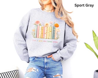 Puede incluir: Una sudadera gris deportivo de cuello redondo con un gráfico de una fila de libros coloridos coronados por varias flores. Debajo de los libros, el texto dice: "Just a girl who loves books". La sudadera tiene mangas largas y puños.