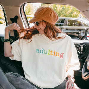 May include: Ivory-colored t-shirt with the word "adulting" in rainbow colors. The person in the image is wearing a corduroy hat and sunglasses. The image is taken inside a car.