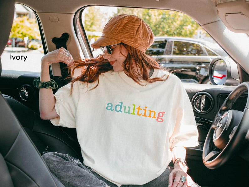 May include: Ivory-colored t-shirt with the word "adulting" in rainbow colors. The person in the image is wearing a corduroy hat and sunglasses. The image is taken inside a car.