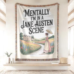 May include: Cream-coloured throw blanket with a printed design featuring the text "Mentally I'm in a Jane Austen Scene." The illustration depicts a woman with an umbrella walking on a path towards a manor house. The blanket has a fringed edge.