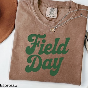 May include: Espresso-colored t-shirt with the words "Field Day" in green script. The shirt has a crew neck and short sleeves. A silver necklace with a small pendant is draped across the shirt. The shirt is made of a soft, comfortable material.