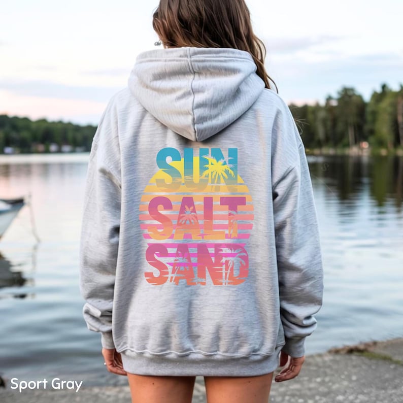 K&ouml;nnte beinhalten: Ein sportgrauer Kapuzenpullover mit einer Grafik auf der R&uuml;ckseite, die den Text SUN SALT SAND in einem Farbverlauf aus Blau, Gelb, Orange und Pink zeigt. Das Design enth&auml;lt Palmensilhouetten und horizontale Linien auf hellgrauem Stoff.