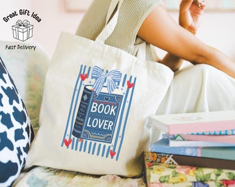 May include: A cream-colored canvas tote bag featuring a blue graphic print of two books with a striped bow on top. The text "BOOK LOVER" is printed in white on the front book cover, surrounded by blue vertical stripes and small red heart accents.