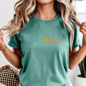 May include: Light green t-shirt with the word "adulting" in rainbow colors. The shirt has short sleeves and a crew neck. The model is wearing the shirt and has blonde hair.