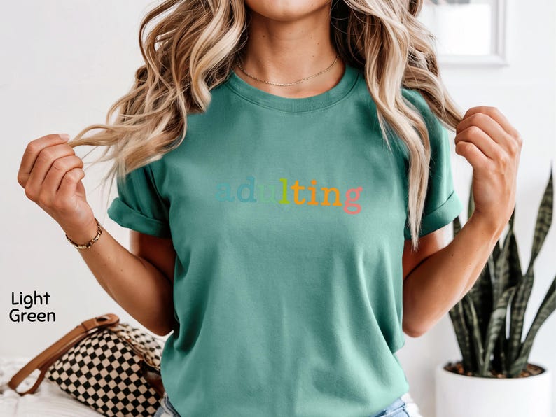 May include: Light green t-shirt with the word "adulting" in rainbow colors. The shirt has short sleeves and a crew neck. The model is wearing the shirt and has blonde hair.
