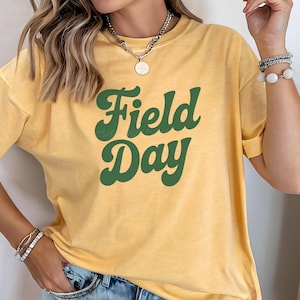 May include: Yellow t-shirt with the words "Field Day" in green, bold, cursive lettering. The shirt has short sleeves and a crew neck. The fabric appears soft and comfortable.