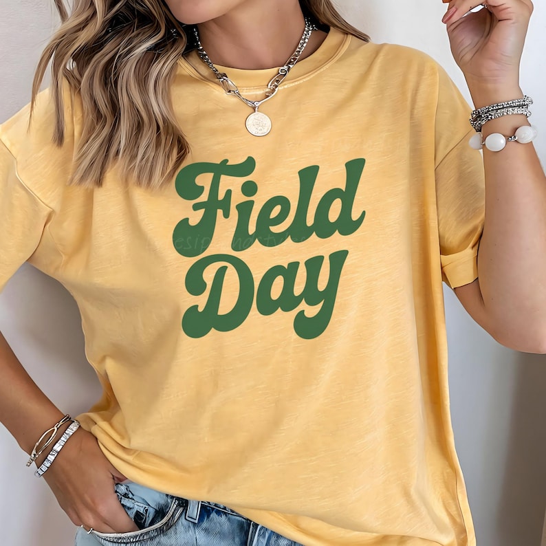 May include: Yellow t-shirt with the words "Field Day" in green, bold, cursive lettering. The shirt has short sleeves and a crew neck. The fabric appears soft and comfortable.