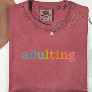 May include: Crimson-colored t-shirt with the word "adulting" in colorful letters. A gold necklace with a star pendant rests on the shirt. A brown hat is visible in the upper left corner. The shirt is size XL.