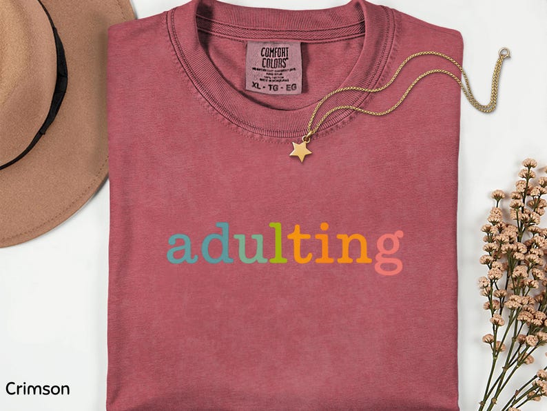 May include: Crimson-colored t-shirt with the word "adulting" in colorful letters. A gold necklace with a star pendant rests on the shirt. A brown hat is visible in the upper left corner. The shirt is size XL.