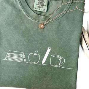 May include: Sage green t-shirt with a white line drawing of books, an apple, a pencil, and a mug. A gold-colored necklace with a small pendant is draped across the shirt. The shirt is folded, and the tag reads "Comfort Colors".