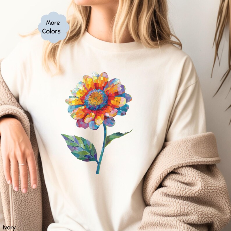 以下が含まれることがあります： アイボリー色の半袖Tシャツ。赤、オレンジ、黄色、青、紫の色合いでモザイク模様のように描かれた大きな花のグラフィックが中央にあります。花には緑色の茎と葉がついています。青い小さなタグには「More Colors」と書かれています。