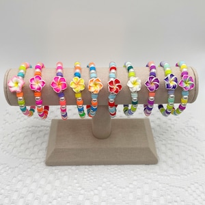Op de afbeelding: Een set van tien kleurrijke armbanden met witte bloemencharms. Elke armband heeft een andere kleurencombinatie van kleine kralen en een witte bloemencharm.