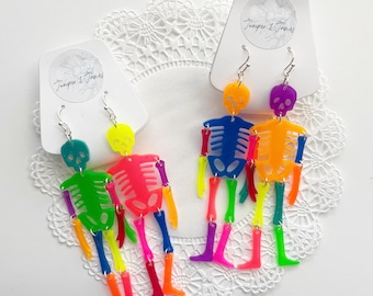May include: A pair of neon skeleton earrings with colorful details. The earrings are made of acrylic and are lightweight and fun to wear.