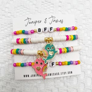 Puede incluir: Cuatro pulseras de cuentas coloridas con las letras "BFF" y dijes de narval. Las pulseras presentan cuentas blancas, arcoíris y doradas. Los dijes son rosas y turquesas con detalles de arcoíris. La tarjeta dice "Juniper & James" y "www.juniperjamesacc.etsy.com".