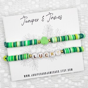 Könnte beinhalten: Zwei grün-weiß gestreifte Armbänder mit goldenen Perlen und einem grünen Kleeblatt-Anhänger. Ein Armband hat das Wort "LUCKY" in goldenen Buchstaben auf weißen Perlen.
