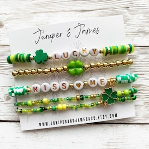 Könnte beinhalten: Vier Armbänder mit grünen und goldenen Perlen, Charms und Buchstaben, die "LUCKY" und "KISS ME" buchstabieren. Die Armbänder befinden sich auf einer weißen Karte mit dem Text "Juniper & James" und einer Website-Adresse.