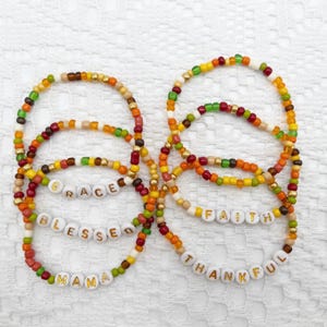Pode incluir: Cinco pulseiras de contas coloridas com as palavras "GRACE", "BLESSED", "MAMA", "FAITH" e "THANKFUL" escritas em letras douradas.