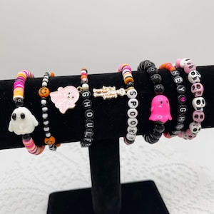 Op de afbeelding: Set van zes Halloween-thema armbanden met bedels. De armbanden zijn gemaakt van zwarte en witte kralen, roze en oranje kralen en zwarte kralen met witte letters. De bedels zijn een spook, een skelet en een hart. De armbanden spellen "Spooky", "Ghoul Life", "Hey Ghoul" en "Boo"