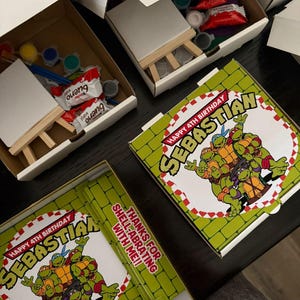 May include: Several open white cardboard boxes containing art supplies and treats. Each box includes a small canvas, a wooden easel, paint pots, and candy. The boxes are decorated with a Teenage Mutant Ninja Turtles theme, with the text "Happy 4th Birthday Sebastian".