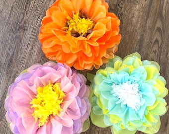 Könnte beinhalten: Drei Seidenpapierblumen in verschiedenen Farben. Die Blumen sind orange, helllila und hellblau mit gelben Akzenten. Die Blumen sind auf einem Holzboden angeordnet.